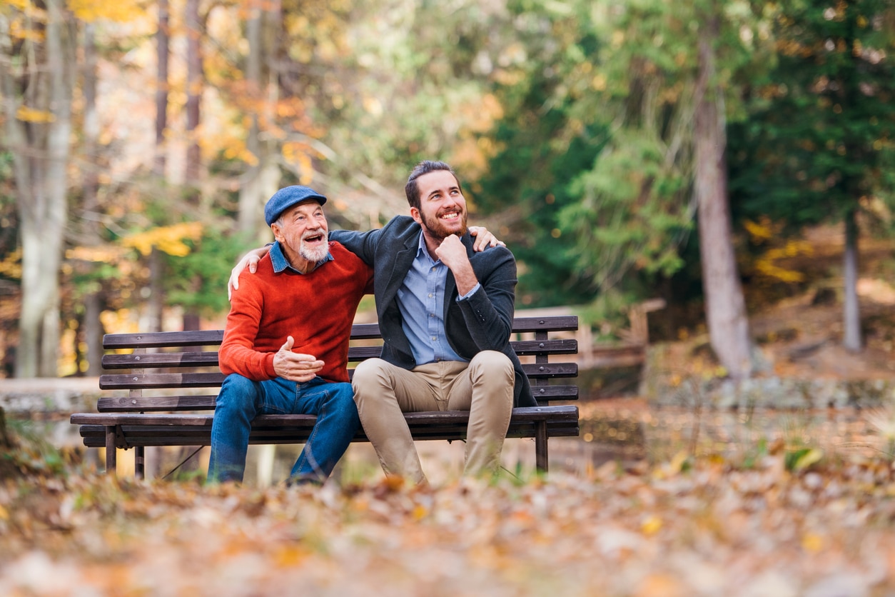 Senior father and his young son sitting on bench by lake in nature, talking.