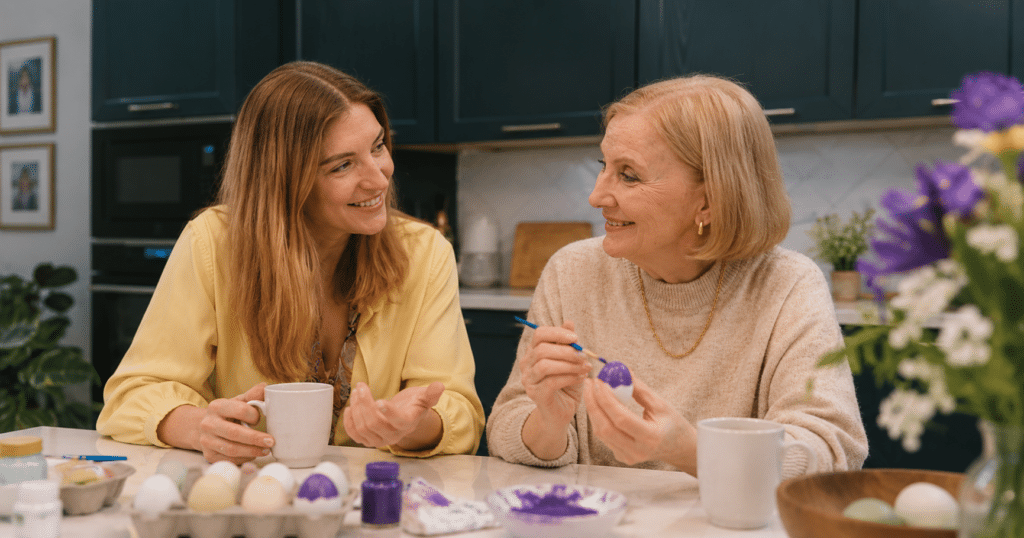 An adult daughter and her older mother talking together at a kitchen table during a relaxed moment at home.