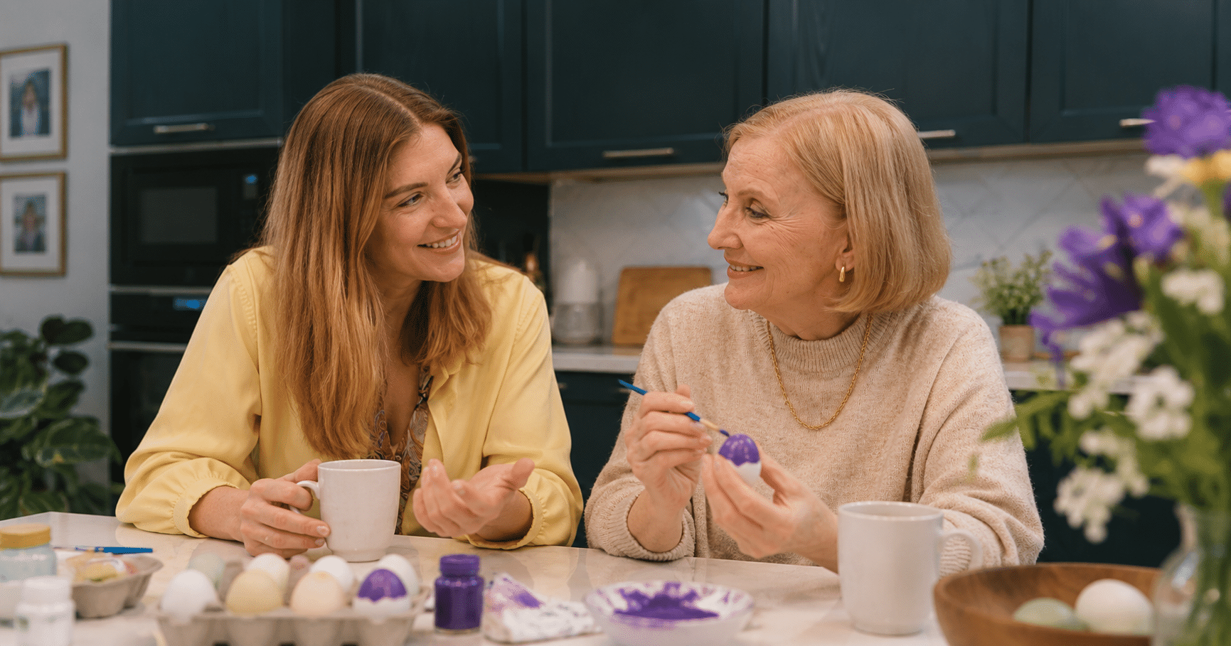 An adult daughter and her older mother talking together at a kitchen table during a relaxed moment at home.
