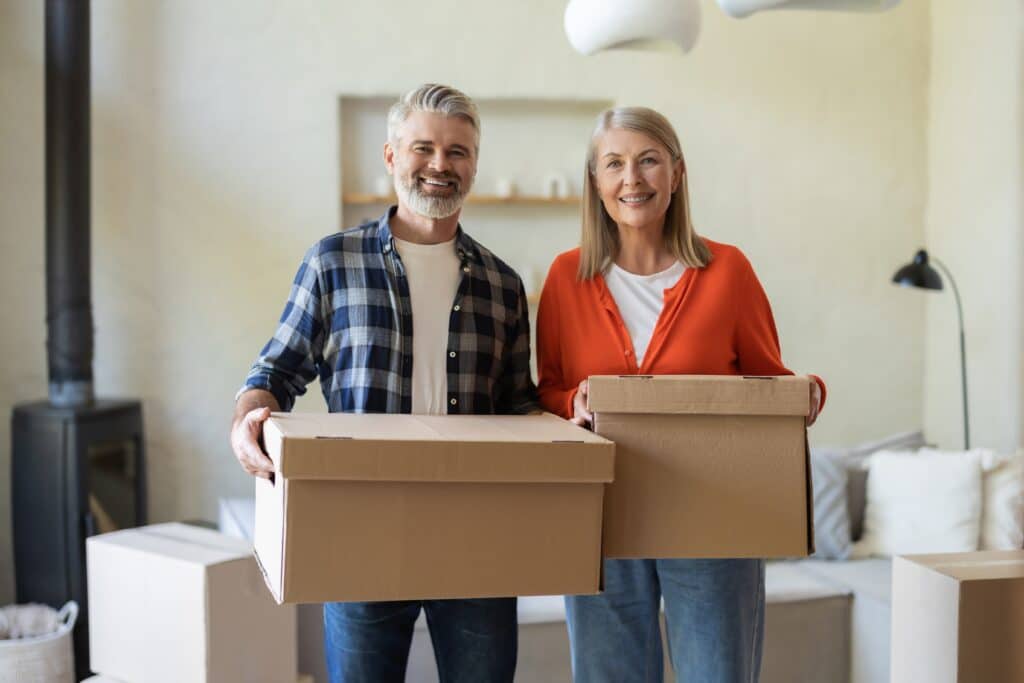 Senior couple holding moving boxes in their home while preparing to downsize, illustrating downsizing for seniors.