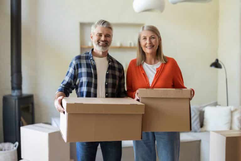 Senior couple holding moving boxes in their home while preparing to downsize, illustrating downsizing for seniors.